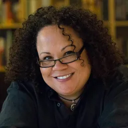 A smiling woman with curly hair and glasses wearing a black collared shirt.