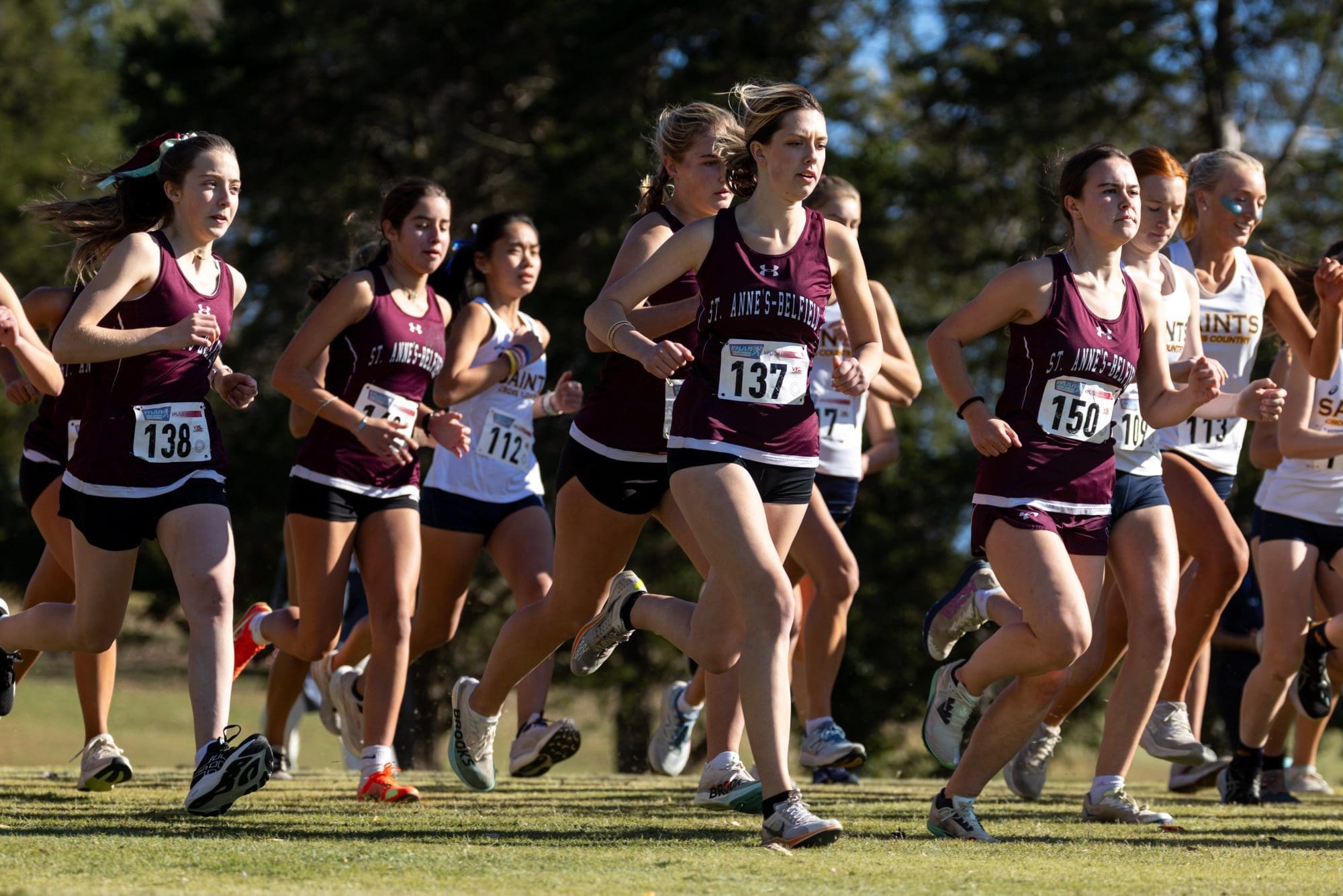 A group of female cross country runners wearing maroon and white uniforms race on a grassy course, with numbered bibs pinned to their shirts, under sunny skies. Trees are visible in the background.