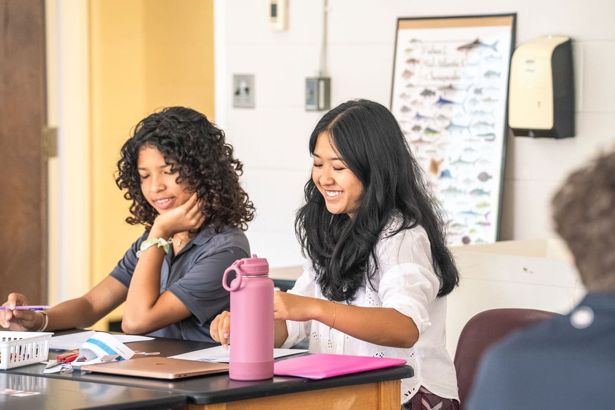 Two students sit at a desk in a classroom, working on papers.