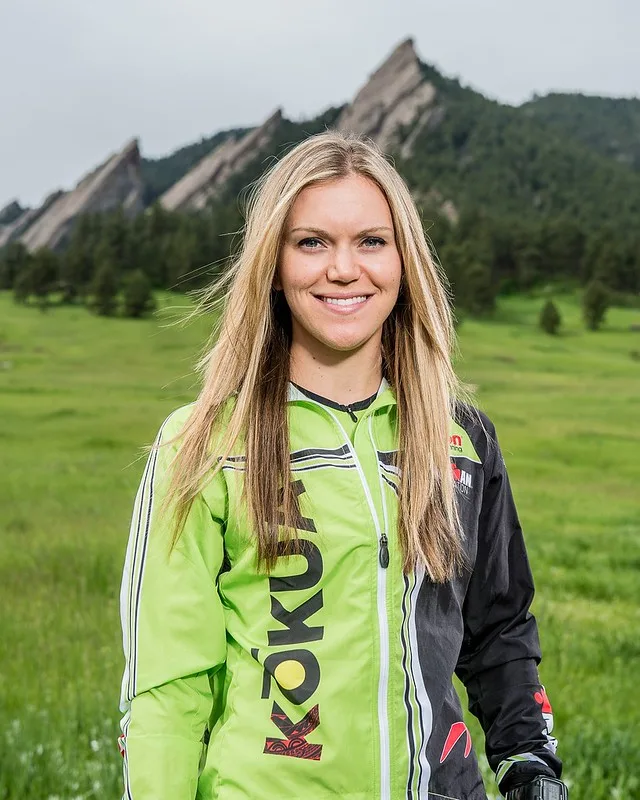 A smiling woman wearing a lime green and black athletic jacket stands in a grassy field with rocky mountain peaks in the background.