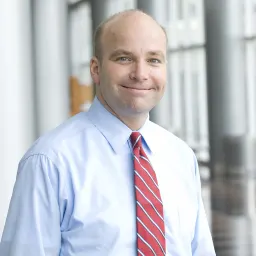A middle-aged man with a receding hairline, wearing a light blue button-down shirt and a red-and-white striped tie.