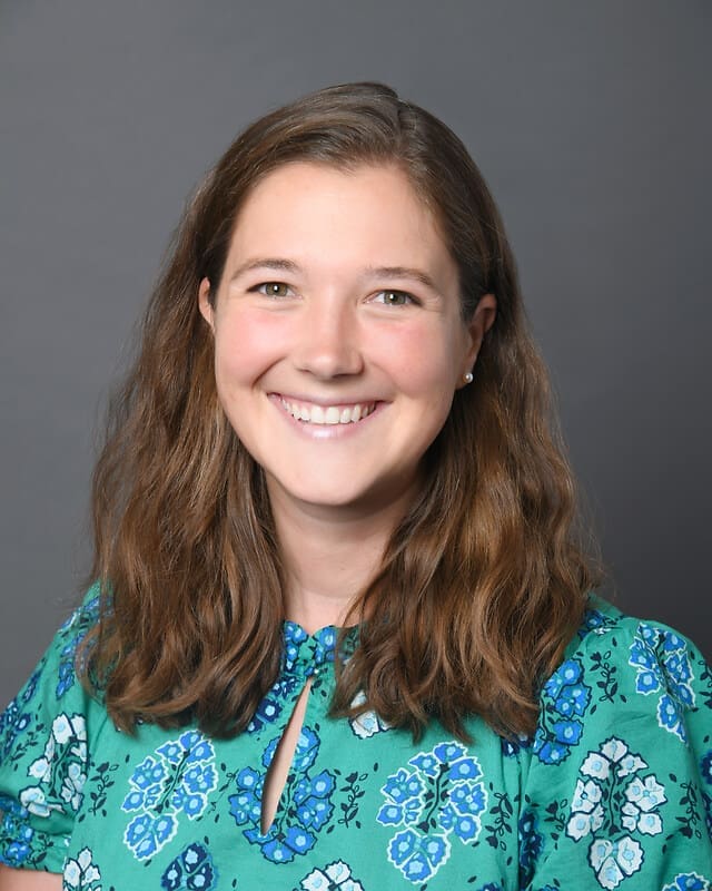 A woman with long brown hair smiles at the camera. She is wearing a green patterned blouse with blue and white designs, and is posed in front of a plain gray background.