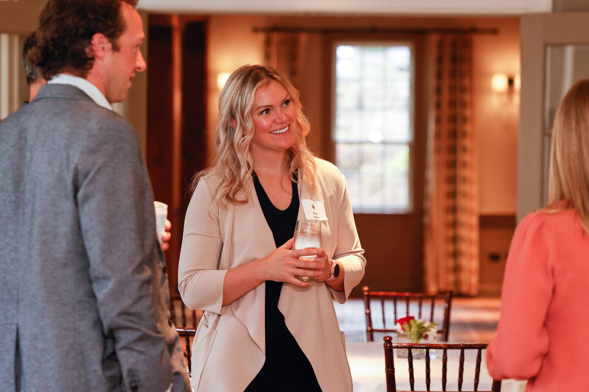 A woman holding a glass and wearing a name tag smiles while talking to two people at a networking event.