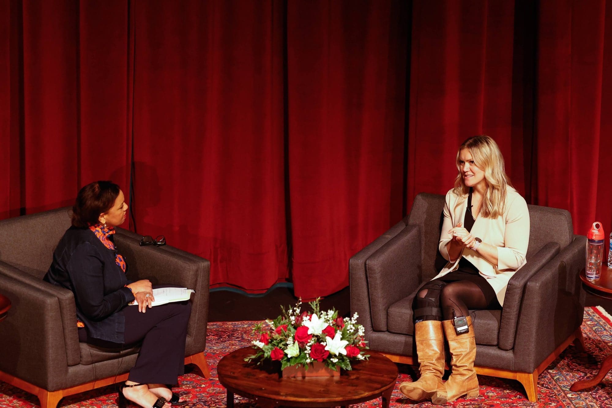 Two women sit in armchairs on a stage with red curtains for an interview, with a flower arrangement on a coffee table between them.