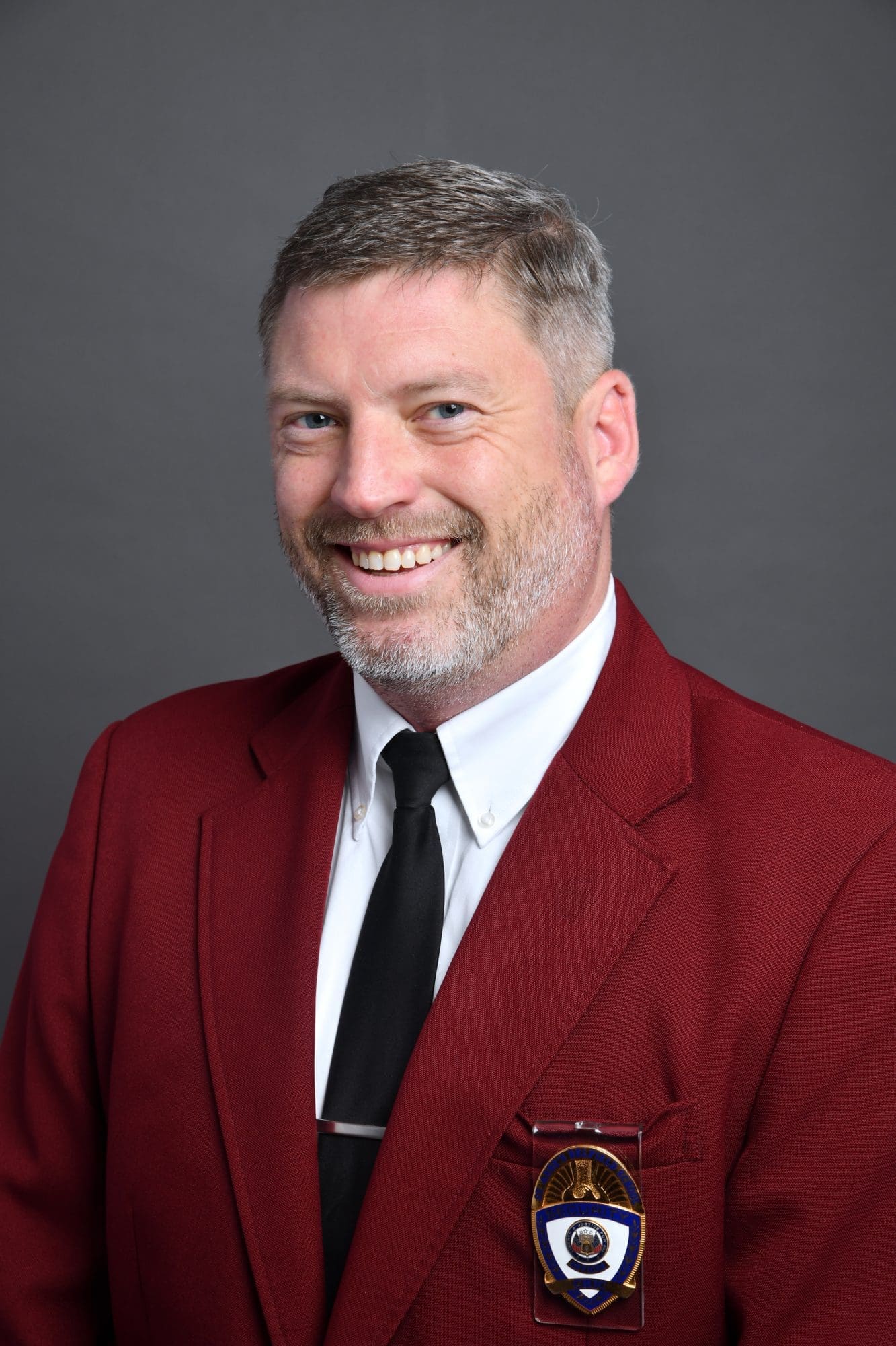 A smiling man with short gray hair and a beard wears a red blazer, white shirt, black tie, and a badge, posing against a plain gray background.