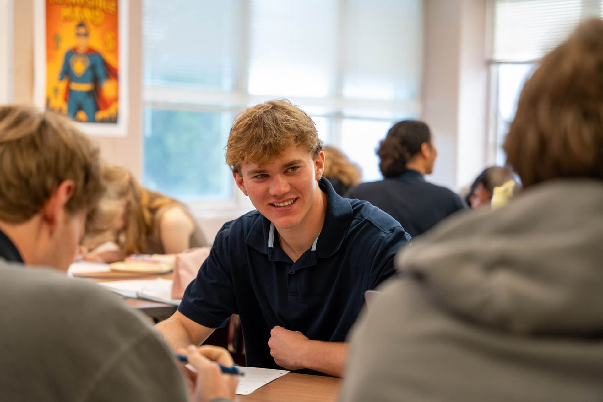 A teenage boy in a navy shirt smiles while sitting at a classroom table with other students. Blurred classmates and a superhero poster are visible in the background.