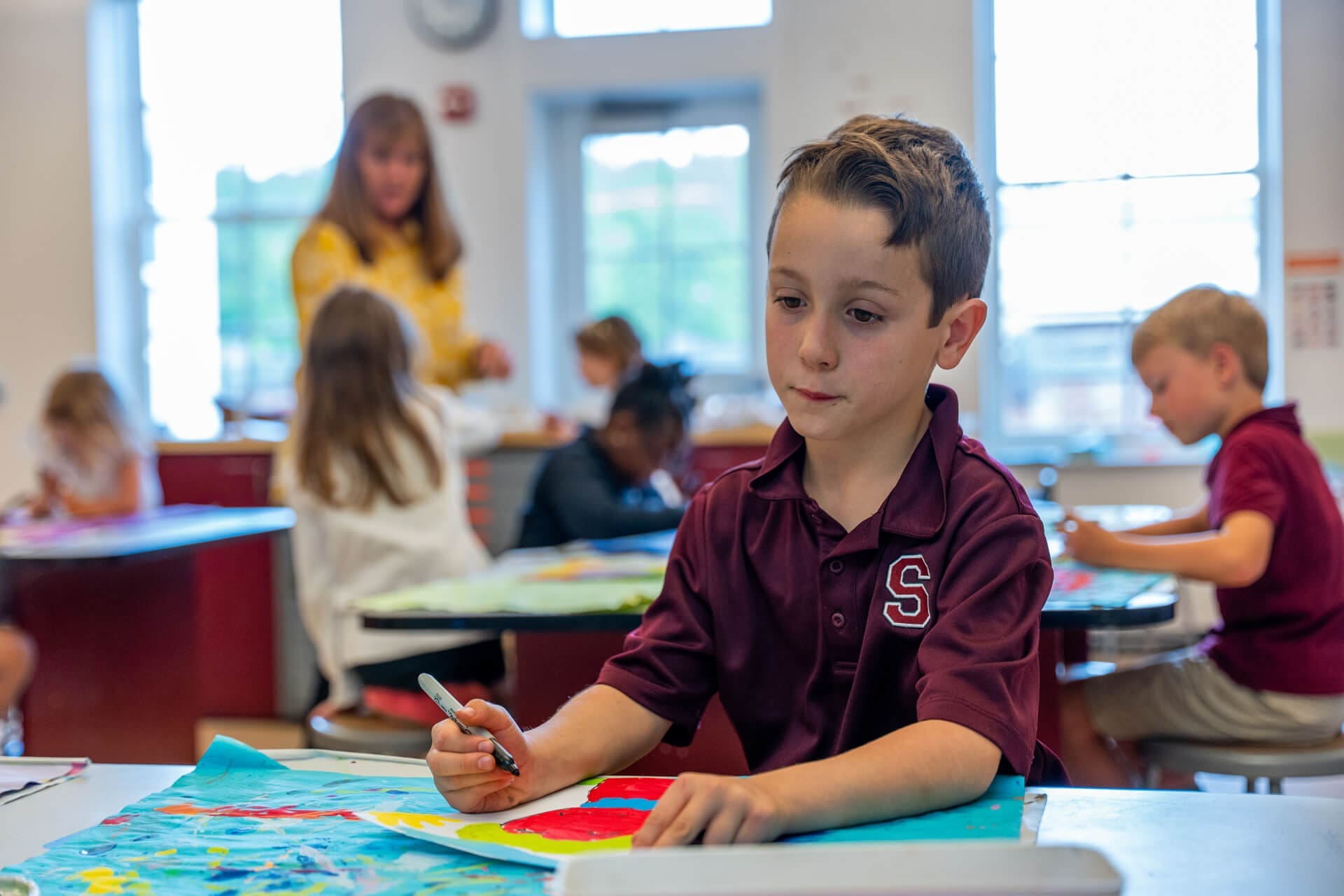 A young boy in a maroon shirt draws with markers at a classroom table. Other children and a teacher are in the background, all engaged in creative activities. The classroom is bright and filled with artwork.