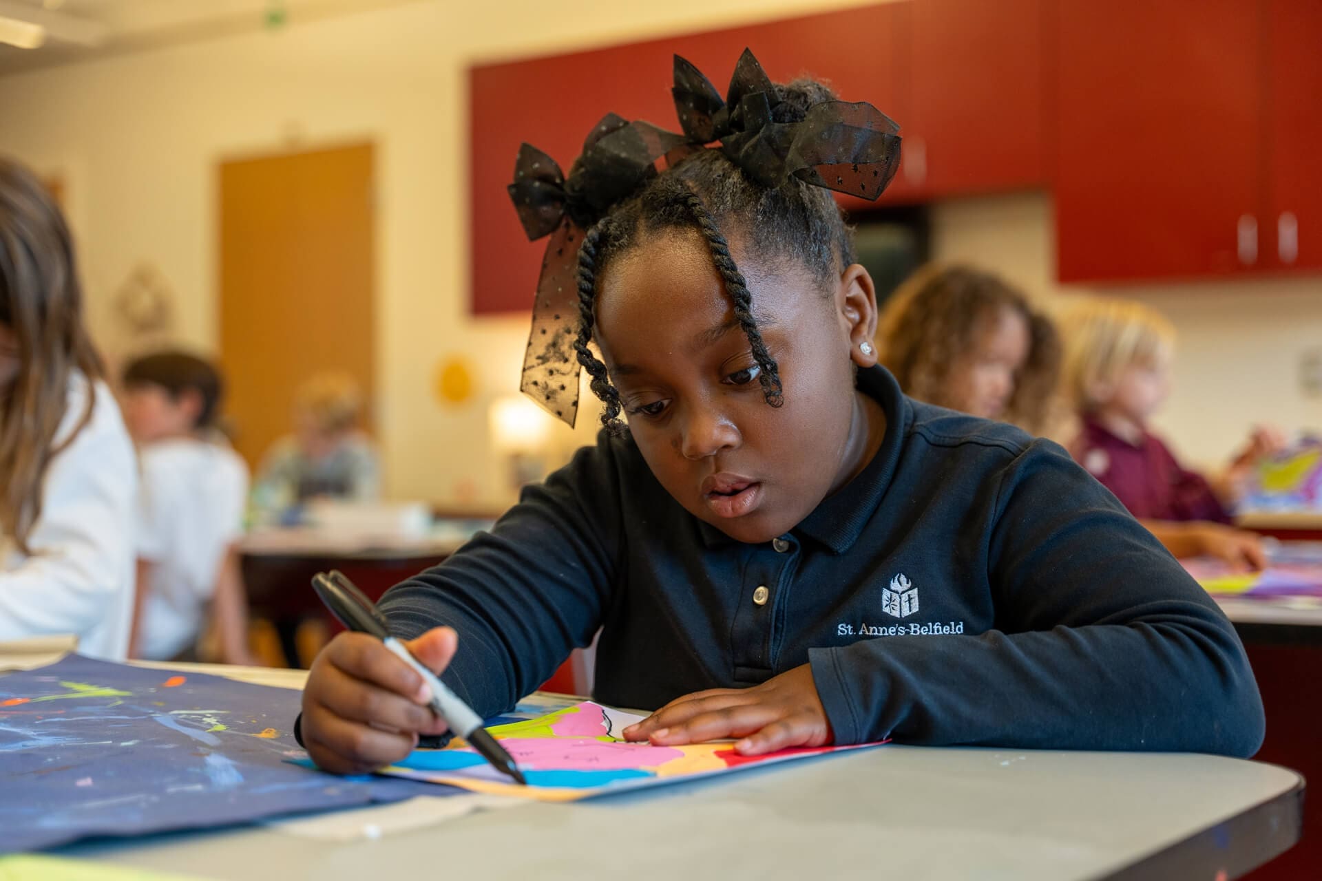A young girl with a black bow in her hair draws with a marker on colorful paper at a classroom table, focused on her artwork. Other children are working in the background.