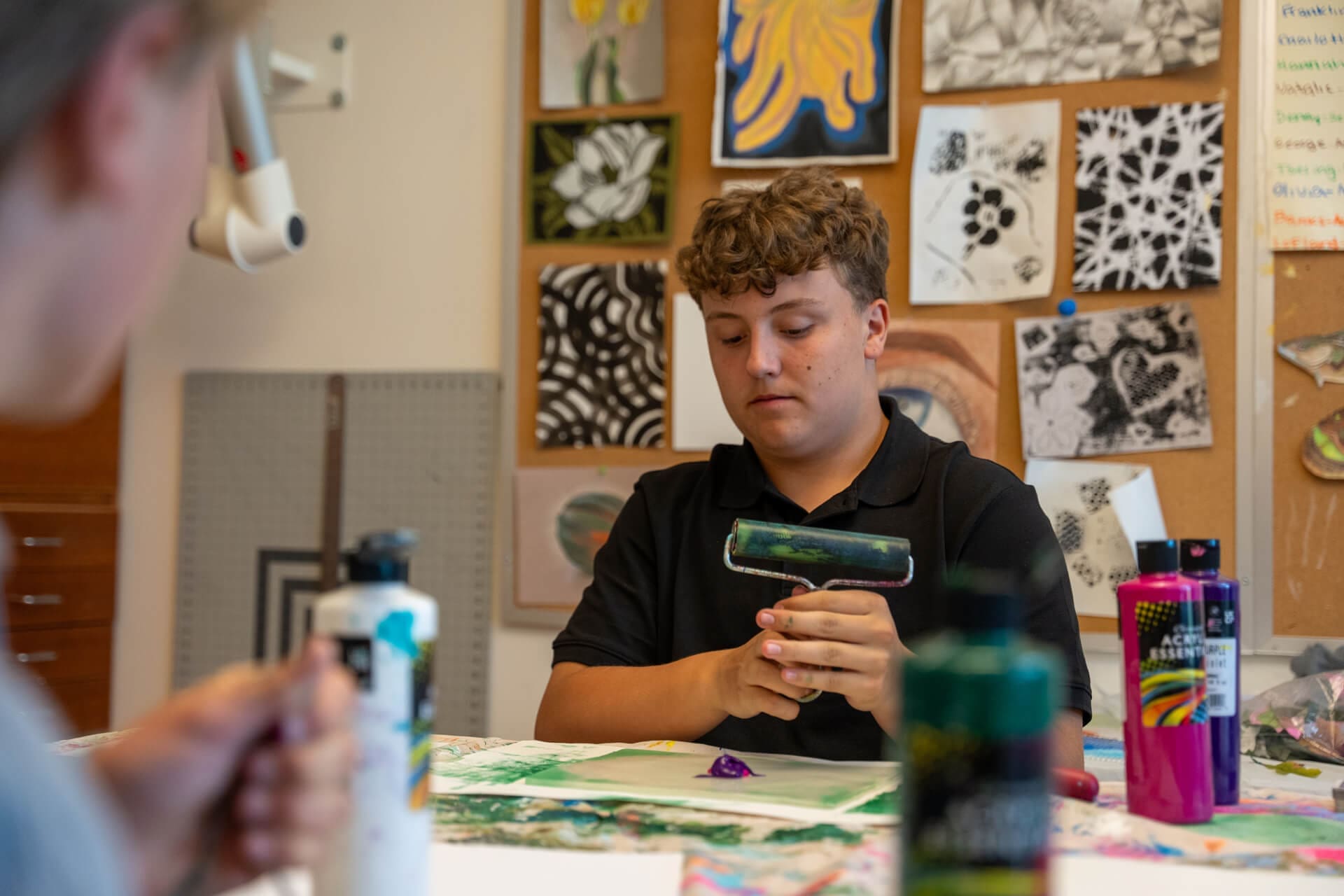 A boy sits at a table in an art classroom, rolling paint onto a brayer. Artworks and drawings are pinned to the wall behind him, while paint bottles and supplies are spread out on the table.