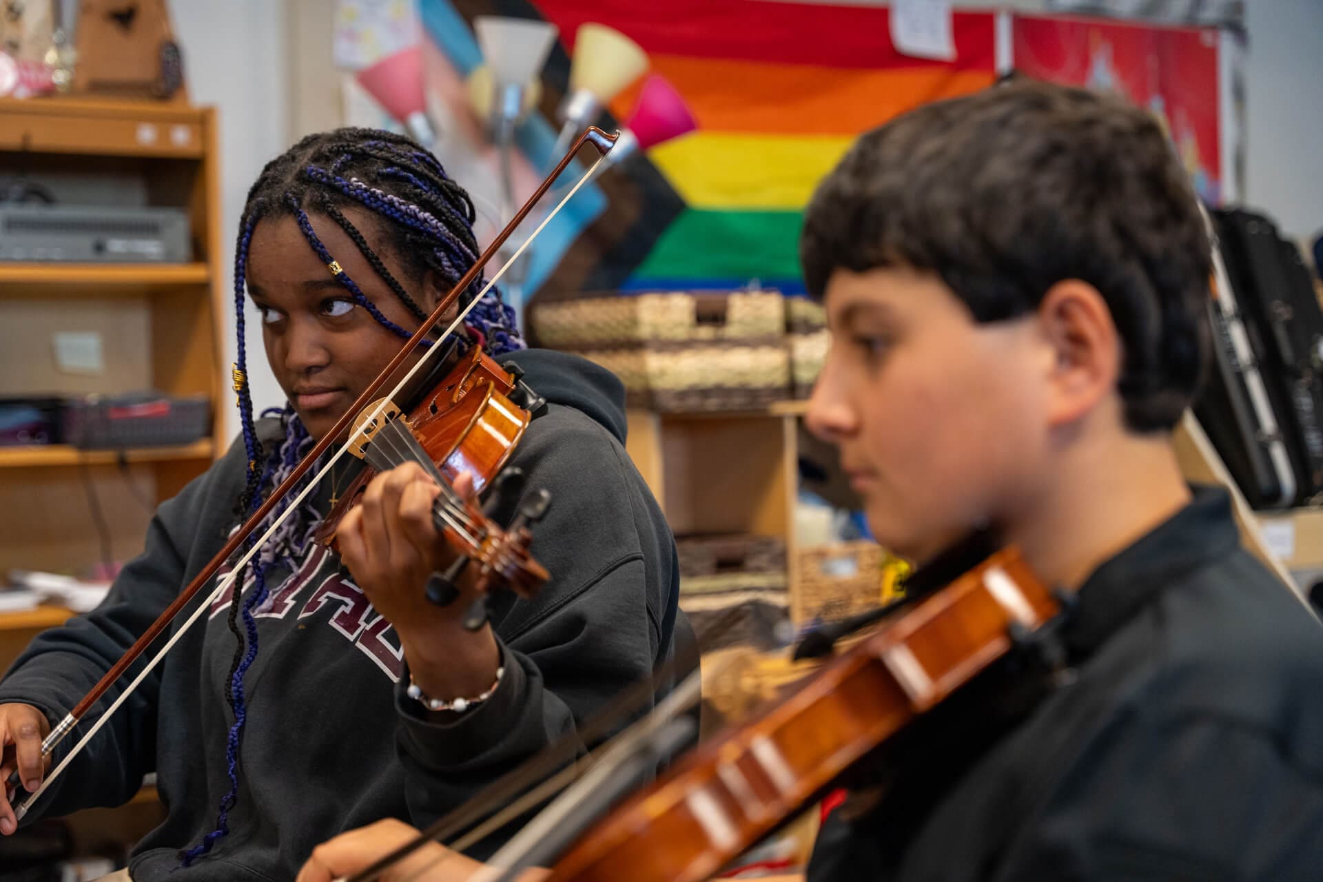 Two students play violins in a classroom. One student with braided hair looks focused, while the other is slightly blurred in the foreground. A rainbow pride flag hangs on the wall in the background.