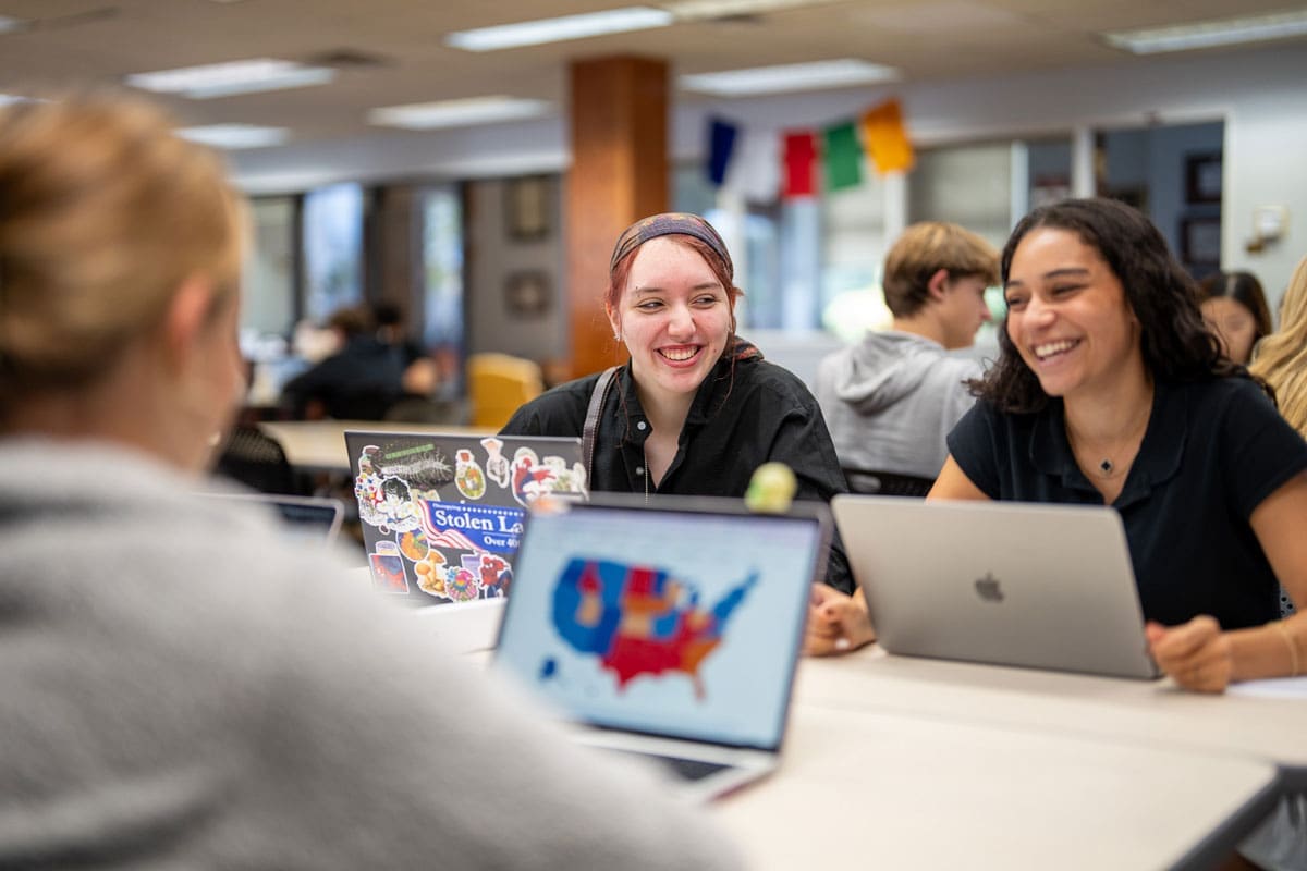 Two female students smiling and talking at a table in a library with laptops open in front of them.