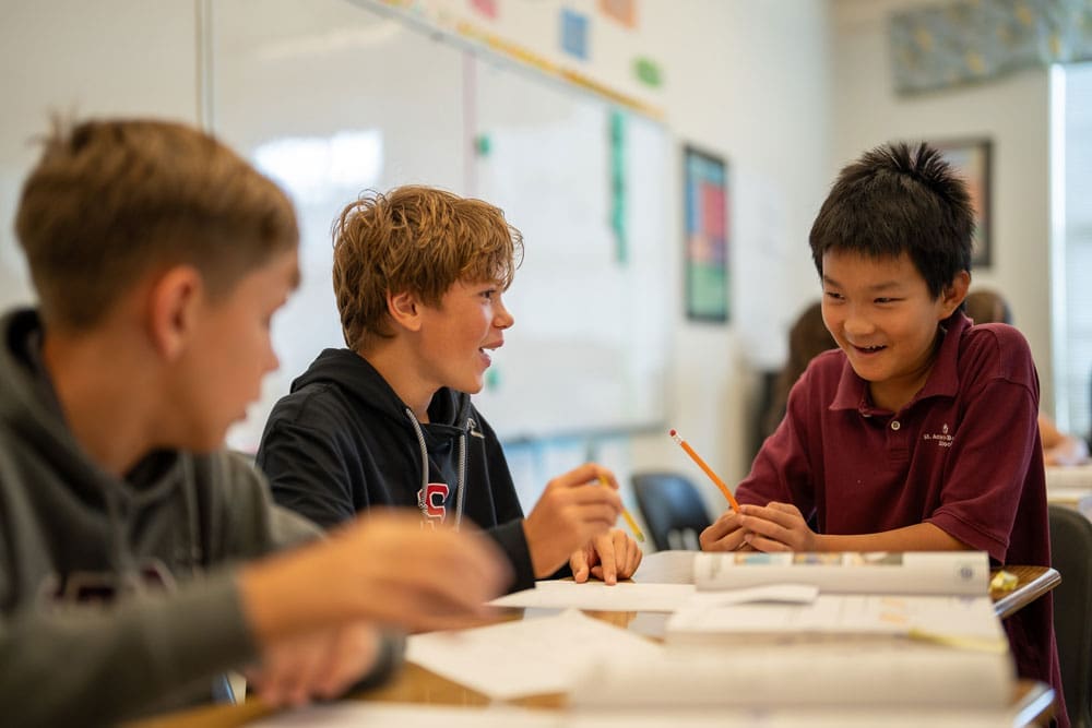 Three students sitting at a classroom desk, talking and working together on an assignment.