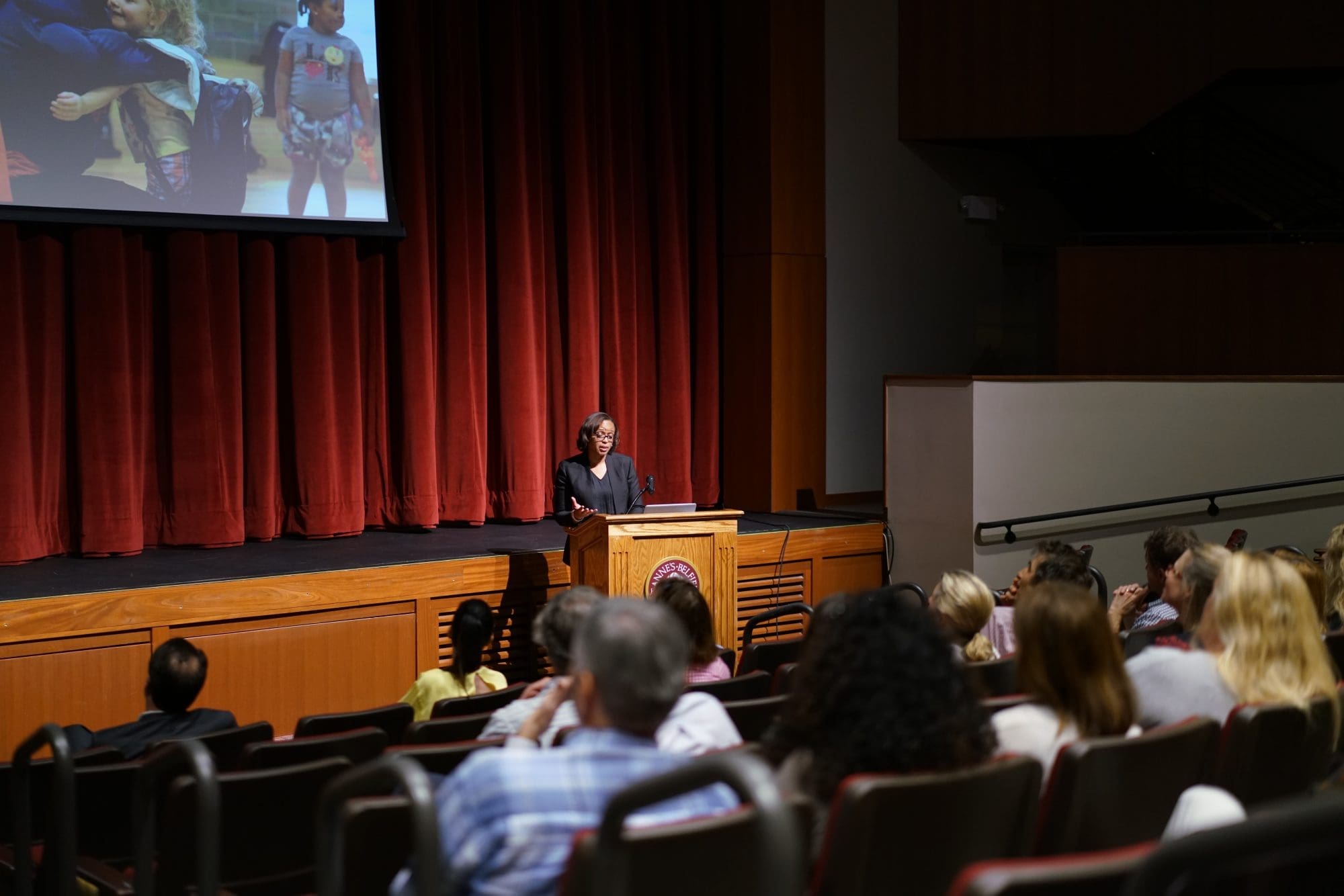 A woman stands at a podium on a stage in front of red curtains as Inspiration Speaks, giving a presentation to an audience seated in a lecture hall.