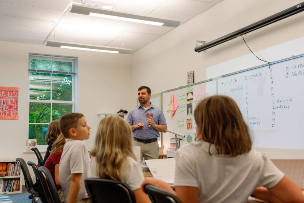 A teacher stands at the front of a classroom speaking to four students seated at desks. The whiteboard behind him displays written notes, and posters decorate the walls. There is a window showing greenery outside.