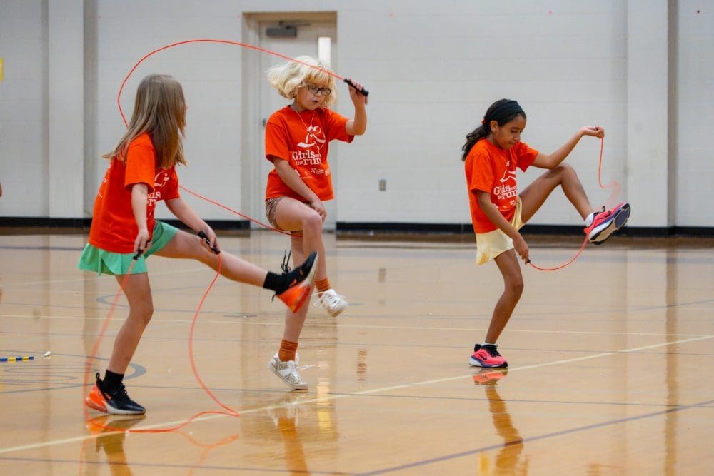 Three girls in matching orange t-shirts jumping rope in a gymnasium.