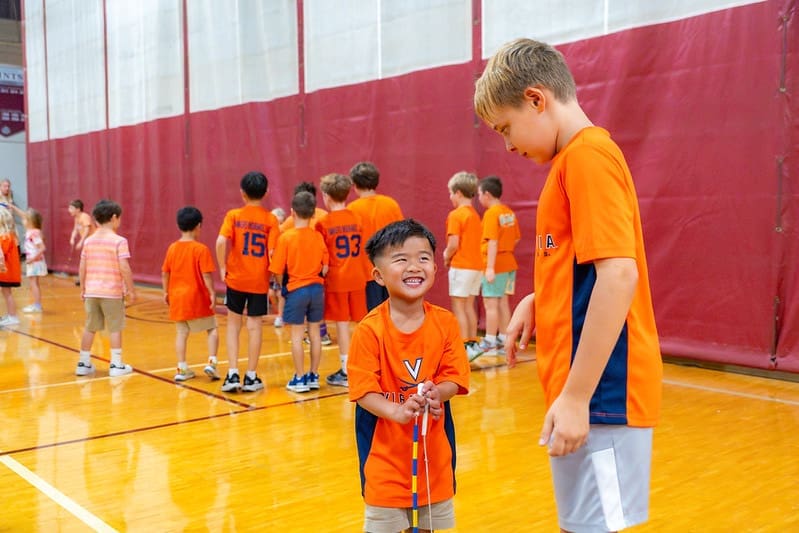 A young boy in an orange sports shirt smiles while holding a jump rope as an older boy watches him on a gymnasium court.
