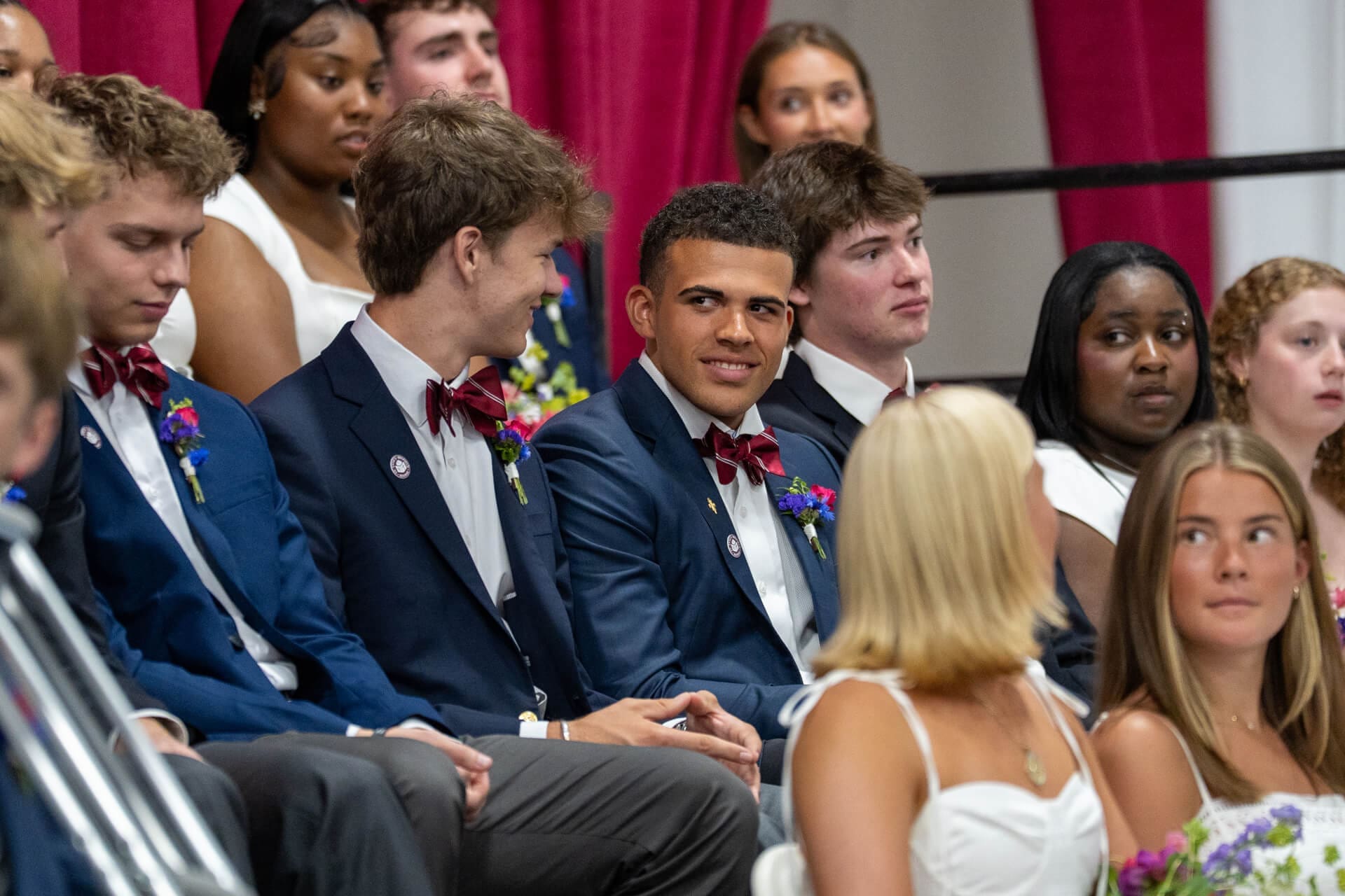 A group of young men in blue suits with red bow ties and young women in white dresses sit together, facing forward, at an indoor event with maroon curtains in the background.