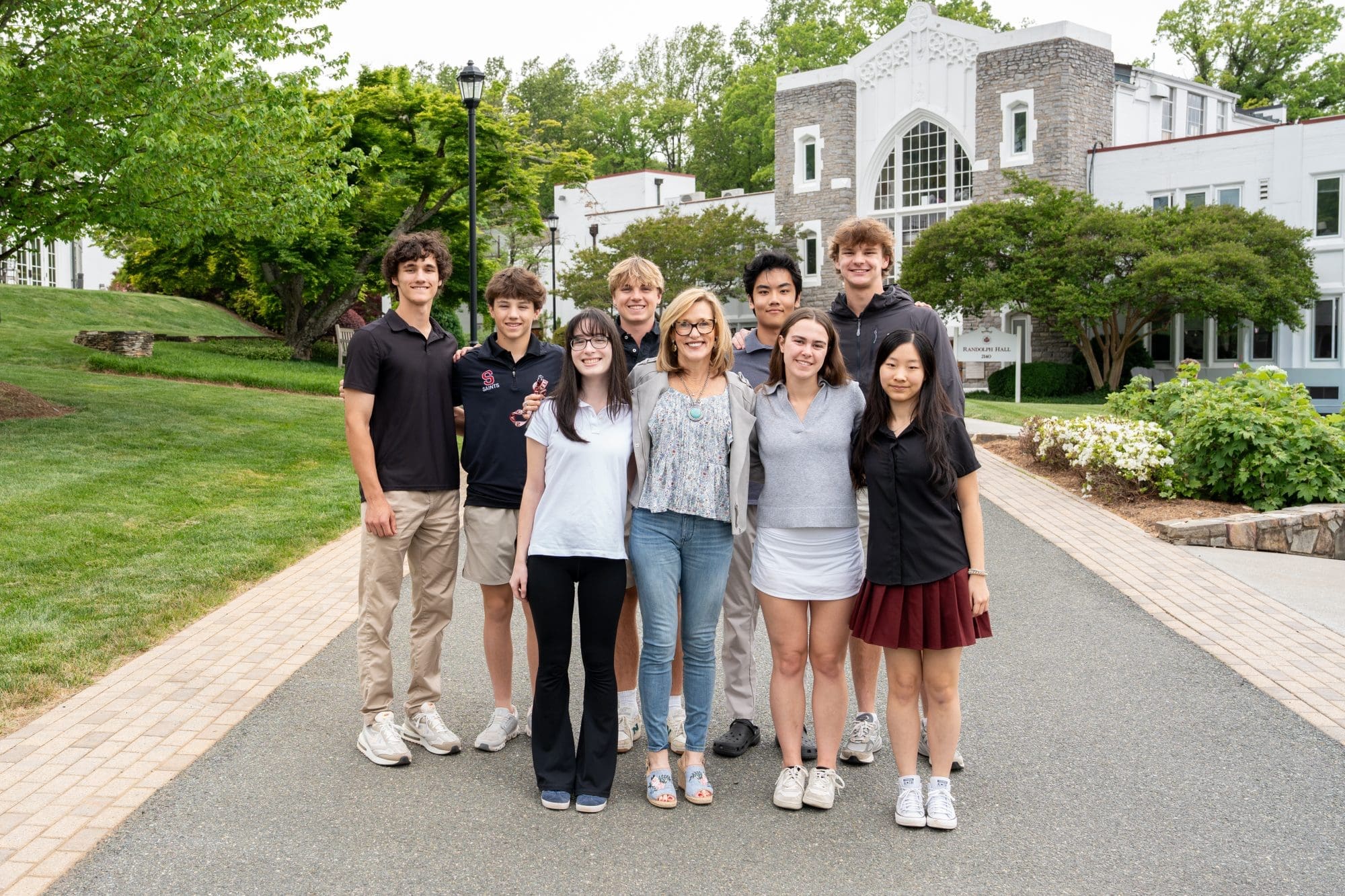 A group of eight young adults and one older adult stand smiling together on a pathway in front of a white building with trees and greenery in the background.