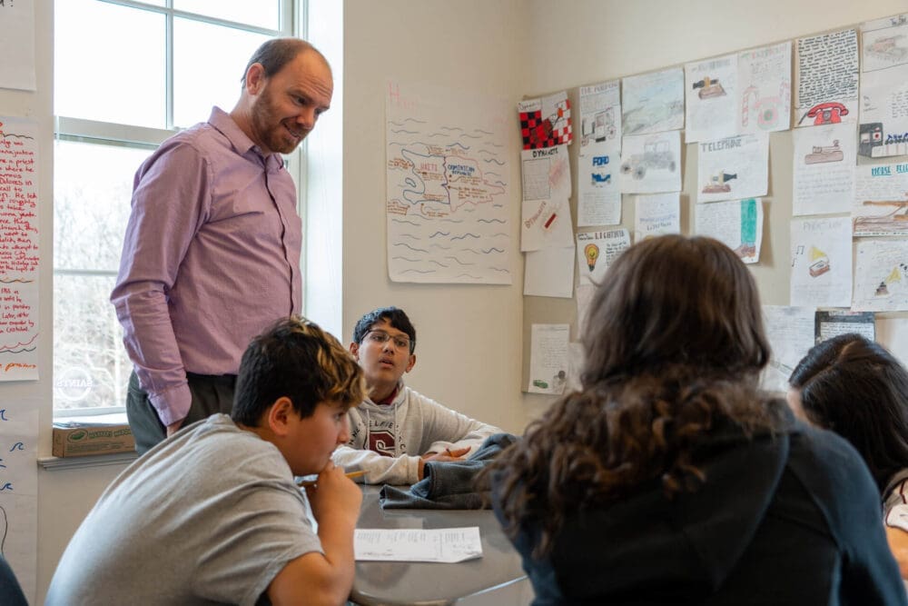 A teacher stands by a group of students seated around a desk in a classroom decorated with posters and drawings on the wall. The students appear to be engaged in discussion.