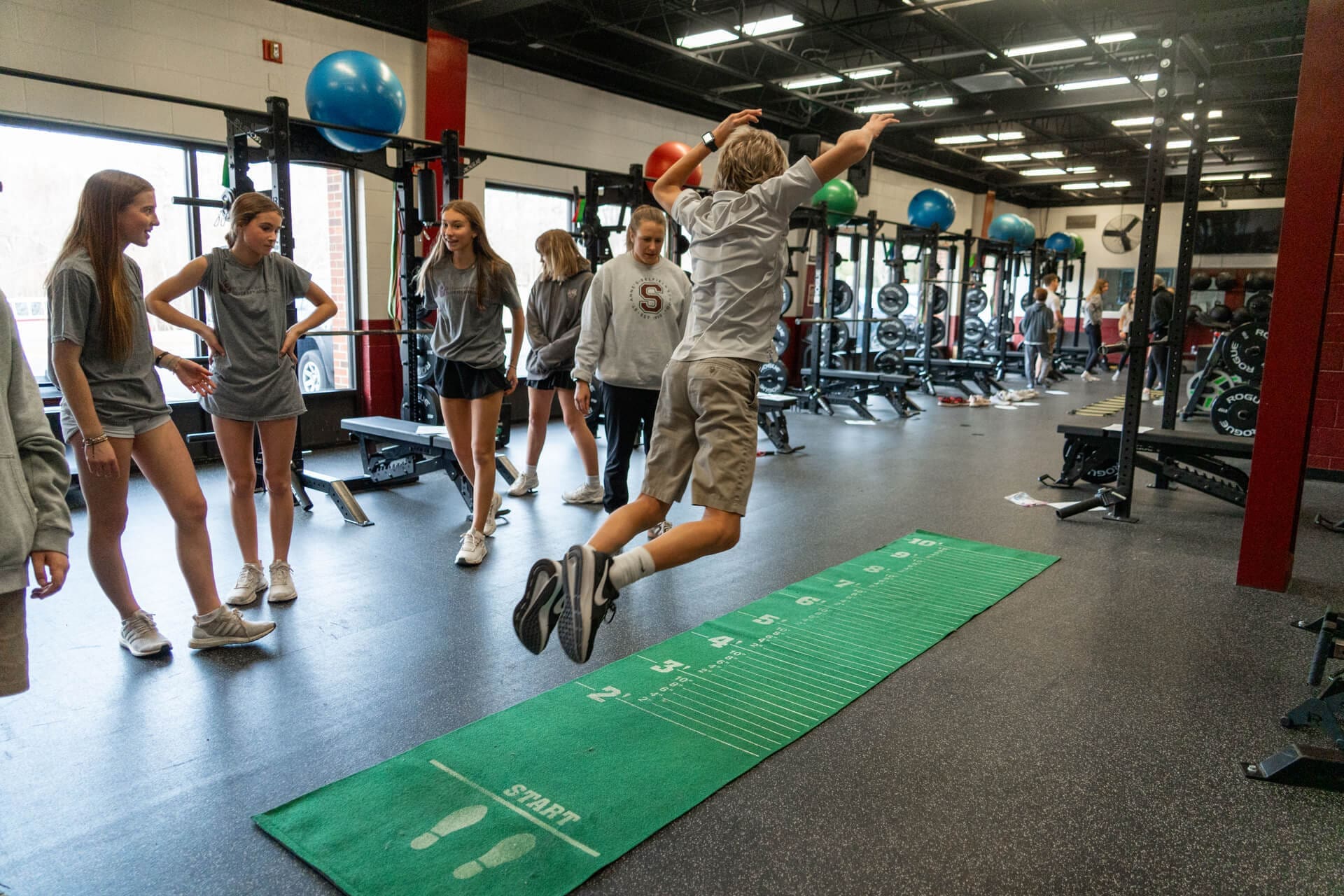 A group of teenagers in athletic wear watch as one boy jumps forward on a green exercise mat marked with measurements in a gym with weights and equipment in the background.