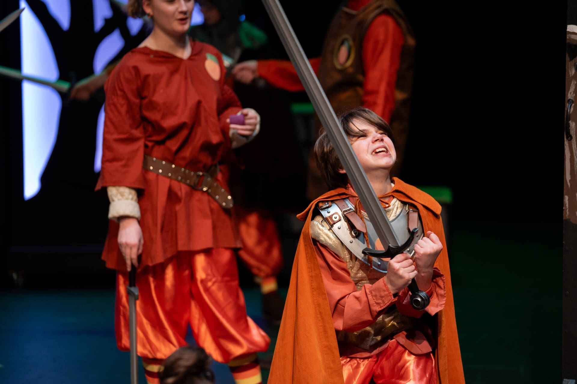 A child in an orange costume and cape holds a large sword with both hands, appearing to shout or strain, while other people in similar costumes stand in the background on stage.