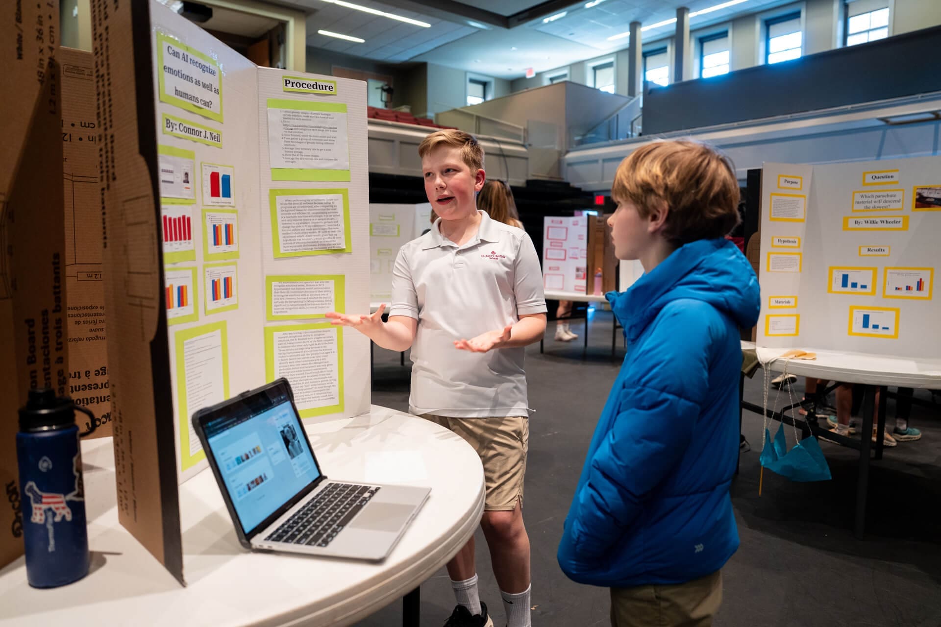 A boy presents his science project to another student at a school science fair, standing by a display board with graphs and text. A laptop and a water bottle are on the table. Other project displays are visible in the background.