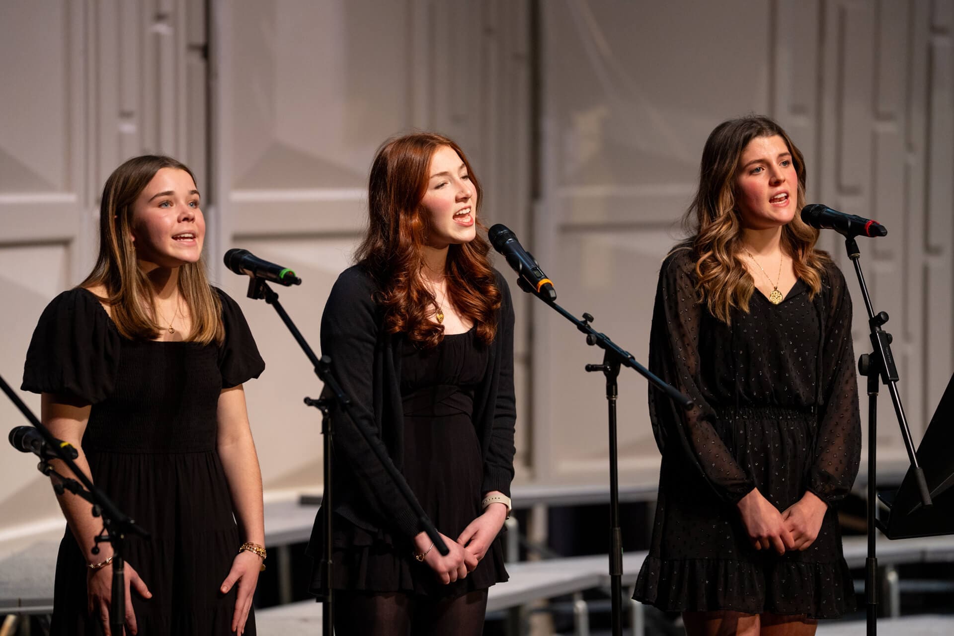 Three young women stand side by side, singing into microphones on stage. They all wear black dresses and are illuminated by stage lights, with a neutral-colored background behind them.