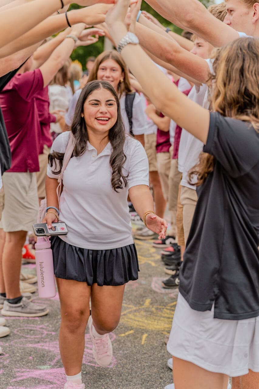 A smiling girl in a white polo and black skirt walks through a tunnel of students at school, giving high fives, holding a pink water bottle and phone, with chalk drawings visible on the ground.