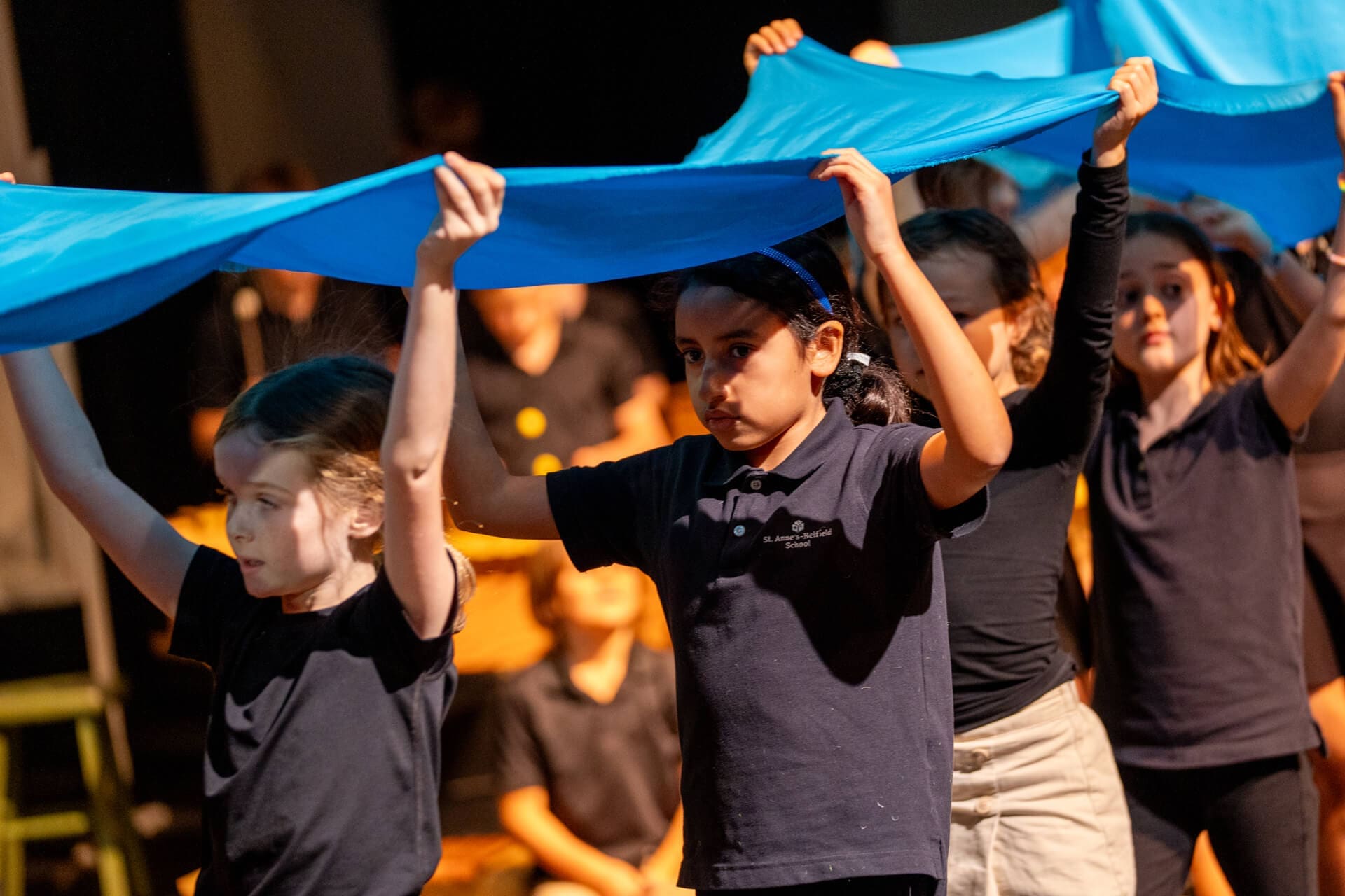 A group of children on stage hold up bright blue fabric above their heads, participating in a performance. They appear focused and are dressed in dark shirts, with warm lighting in the background.