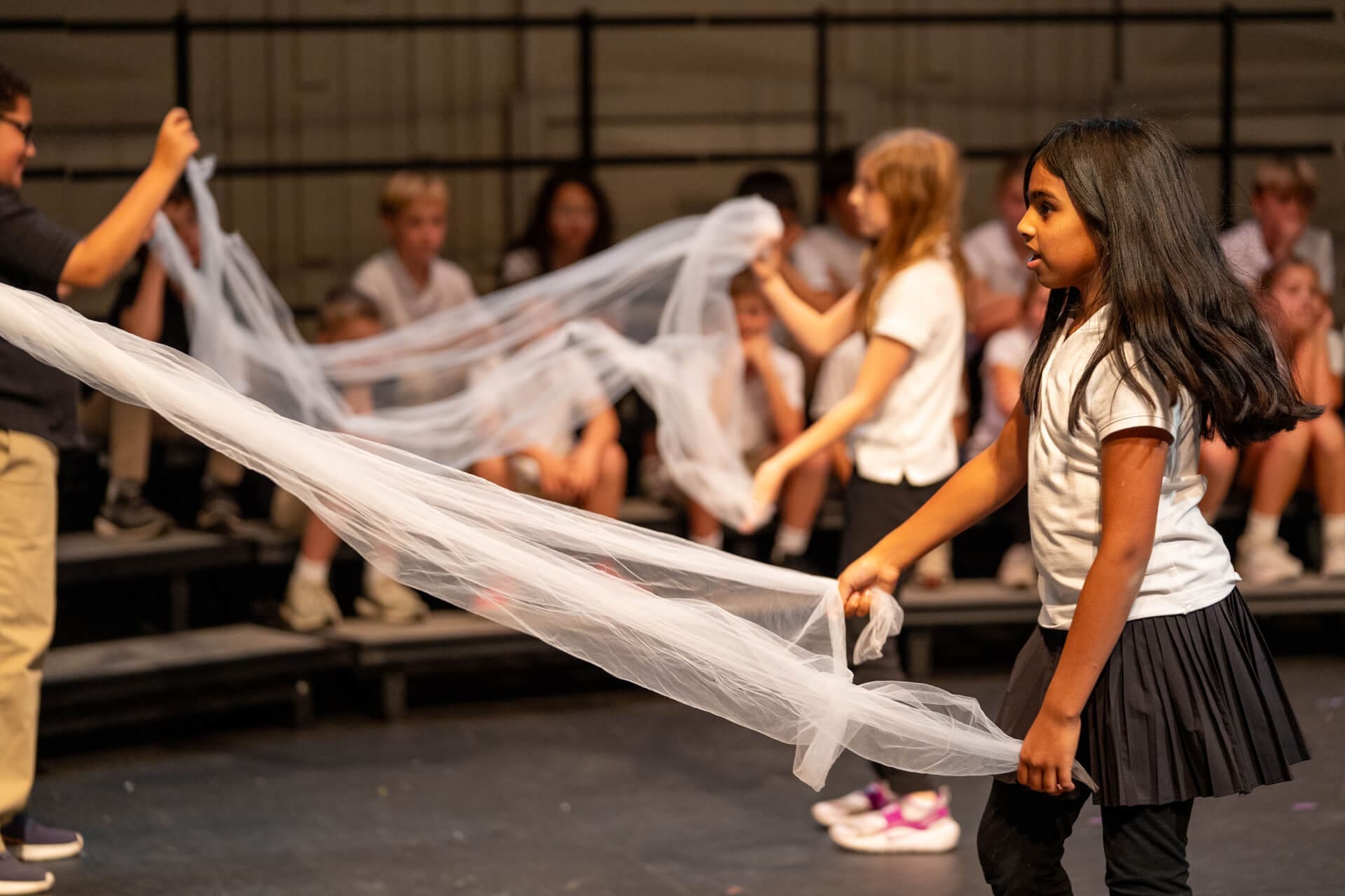 Children stand on stage holding a long, sheer white fabric. One girl in the foreground holds the fabric, while others interact in the background. The setting appears to be a school performance or rehearsal.