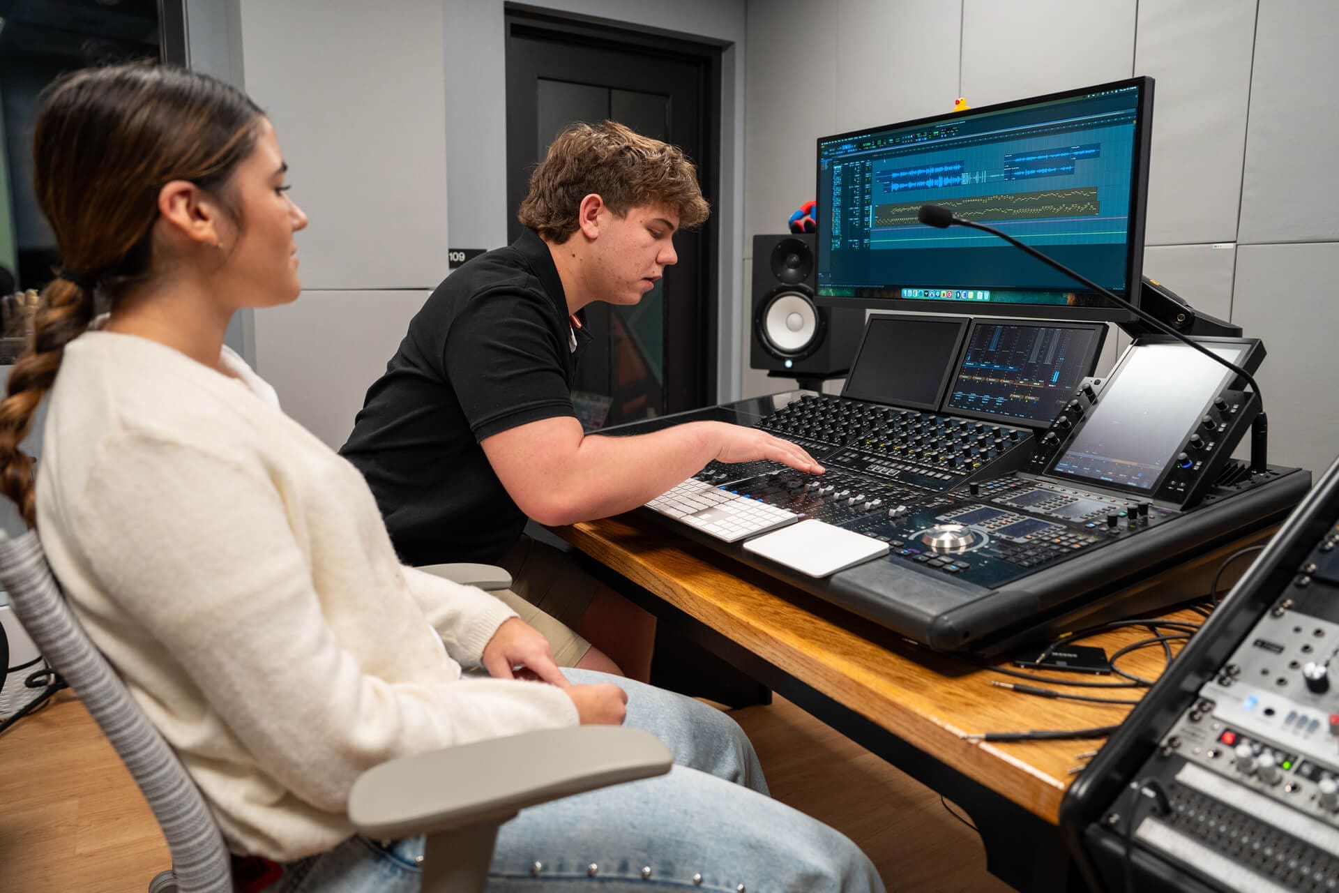 Two people sit in a recording studio; one operates a digital audio mixing console while the other observes. A large monitor displays audio tracks and editing software in a modern, professional school setting.