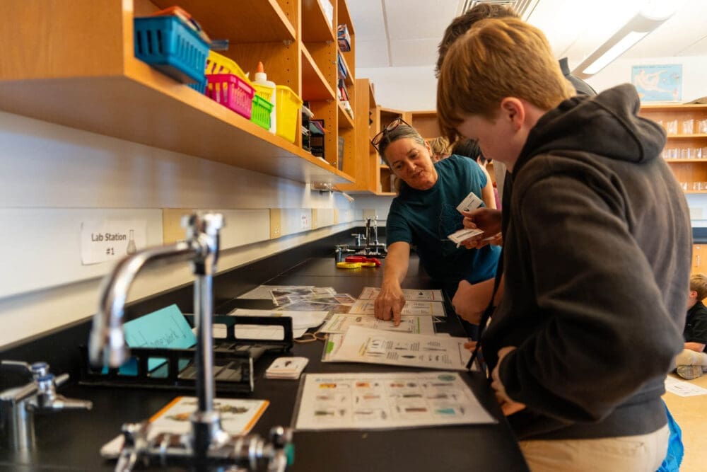 A teacher points at a worksheet on a science lab table, explaining something to two students. The table is covered with papers, lab tools, and bins, and shelves with colorful supplies line the wall.