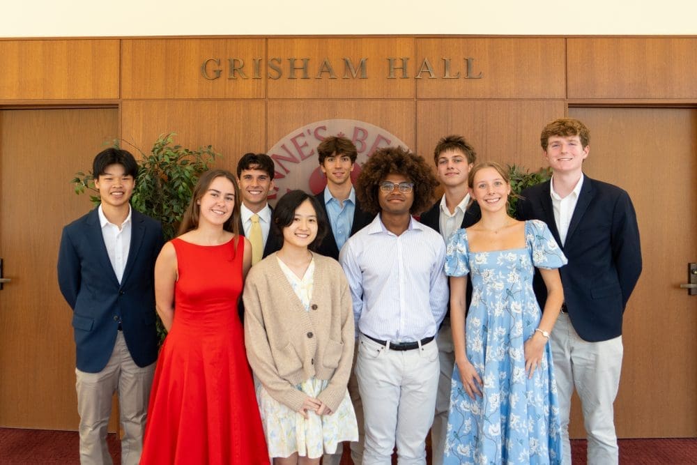 Nine young adults dressed in semi-formal attire smile and pose for a group photo in front of wooden doors labeled GRISHAM HALL with greenery on either side.