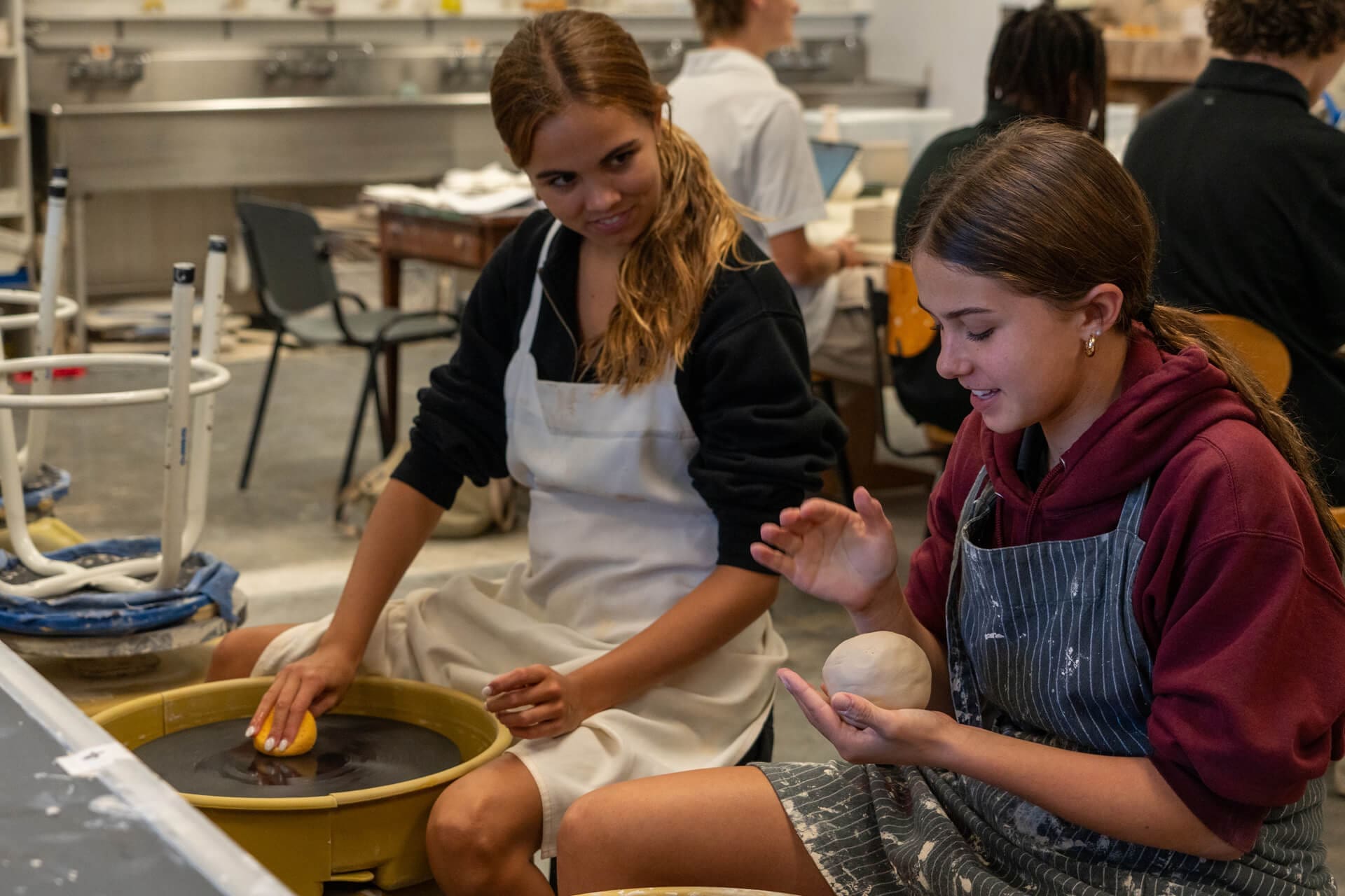 Two young women in aprons work with clay in a pottery studio; one uses a pottery wheel while the other shapes clay with her hands. Other people are seen working in the background.