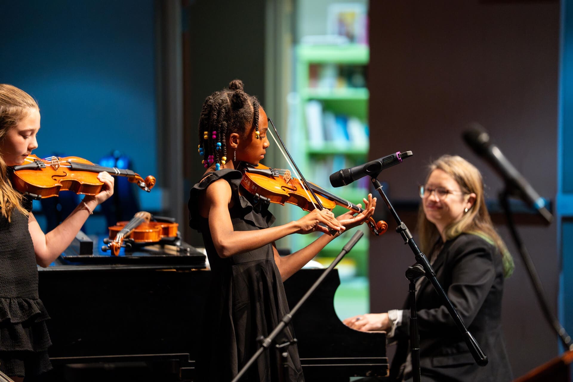 Two young girls play violins on stage, with one girl in the center focused on her instrument. A woman at a piano smiles at them, and microphones are set up in the foreground. Bookshelves are visible in the background.