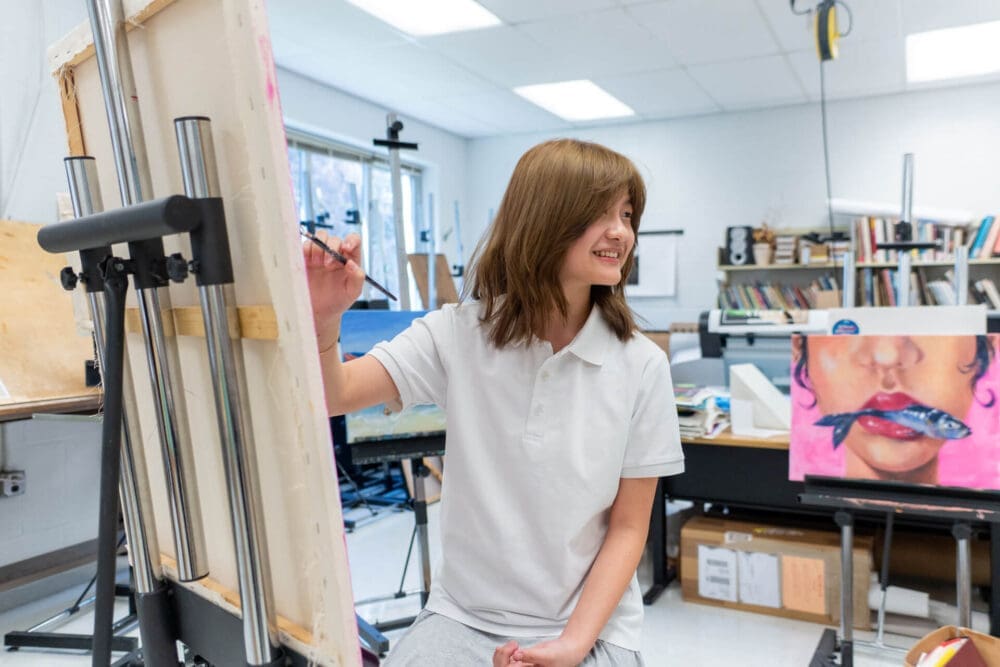 A smiling young person in a white shirt paints on a large canvas in a bright art classroom. Nearby, an easel displays a colorful painting of lips holding a paintbrush. Art supplies and books fill the background.
