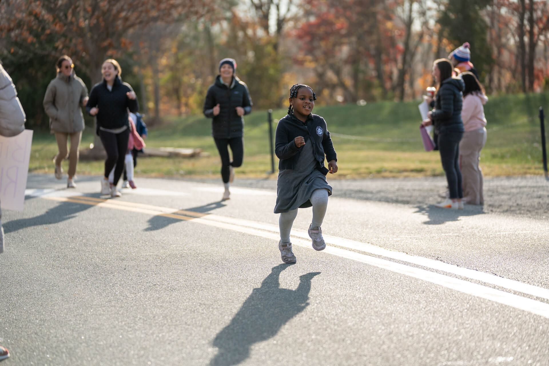 A young girl smiles while running on a road during a race, with other children and adults running behind her and several people standing on the roadside watching and cheering.