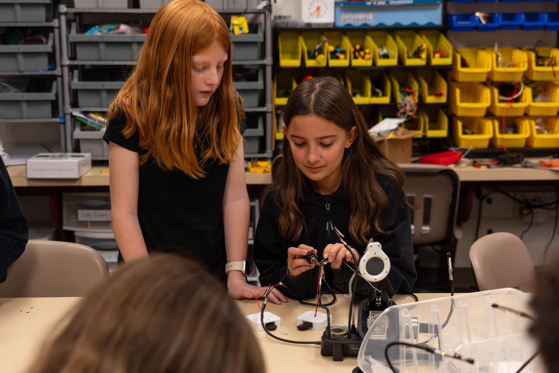 Two girls in a classroom work together on an electronics project at a table, surrounded by tools and bins filled with wires and parts. One girl holds wires while the other observes and assists.