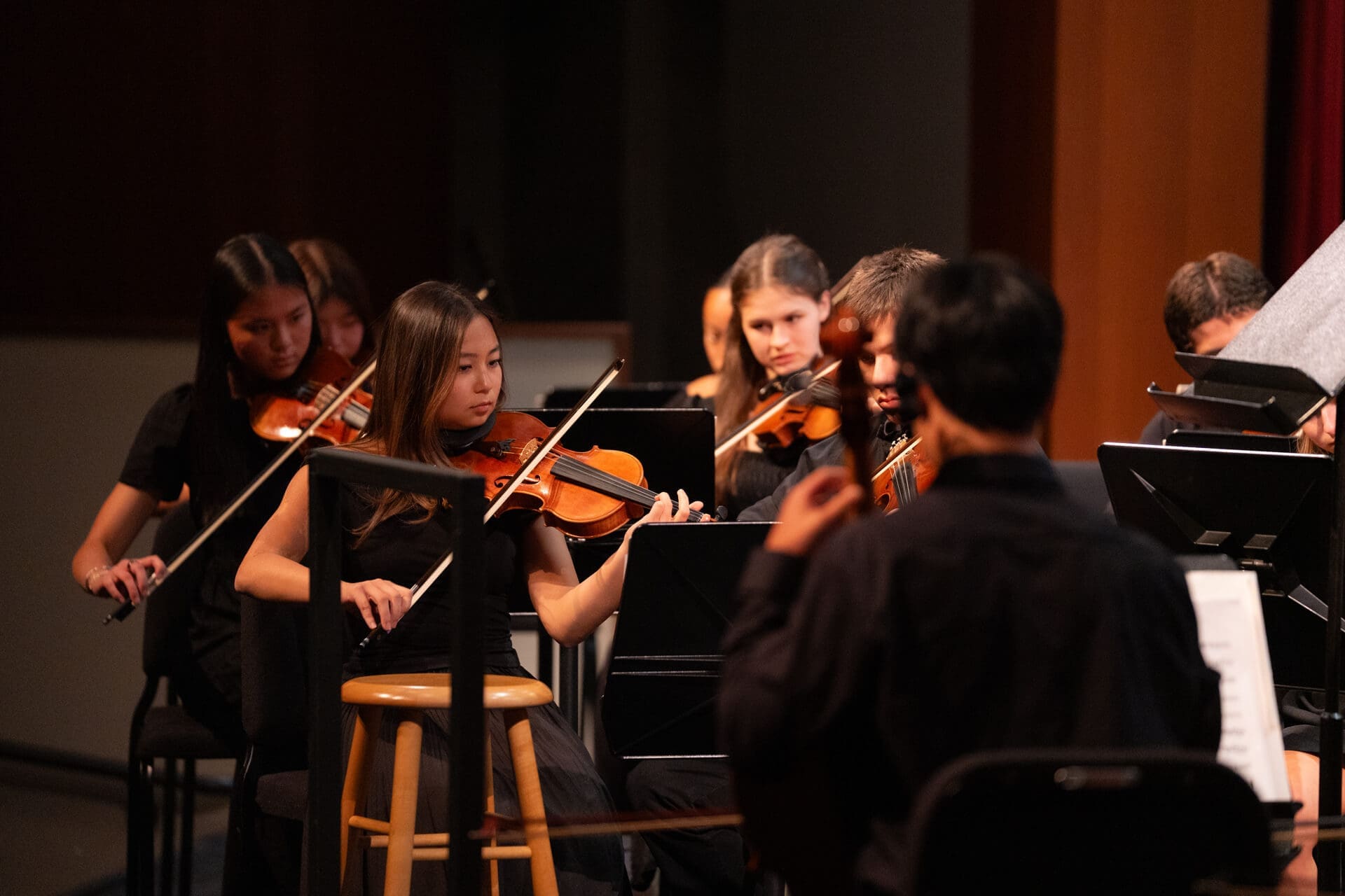 A group of musicians, mostly young women, play violins and violas in an orchestra. They are seated on stage, focused on their music, and dressed in black performance attire.