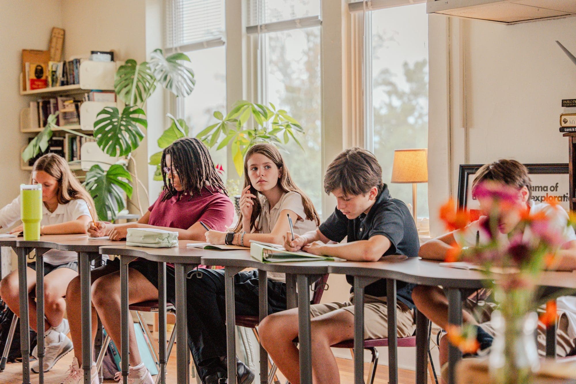 A group of students sits at desks in a bright classroom, writing in notebooks. Large windows and green plants are visible in the background, along with colorful flowers in the foreground.