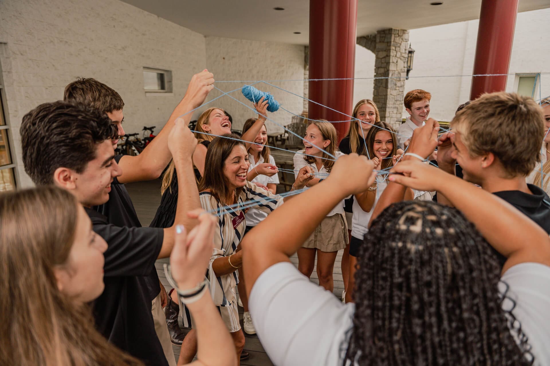 A group of teenagers stands in a circle outside, laughing and holding colorful strings that connect their hands, creating a web in the center. They appear to be participating in a fun team-building activity.