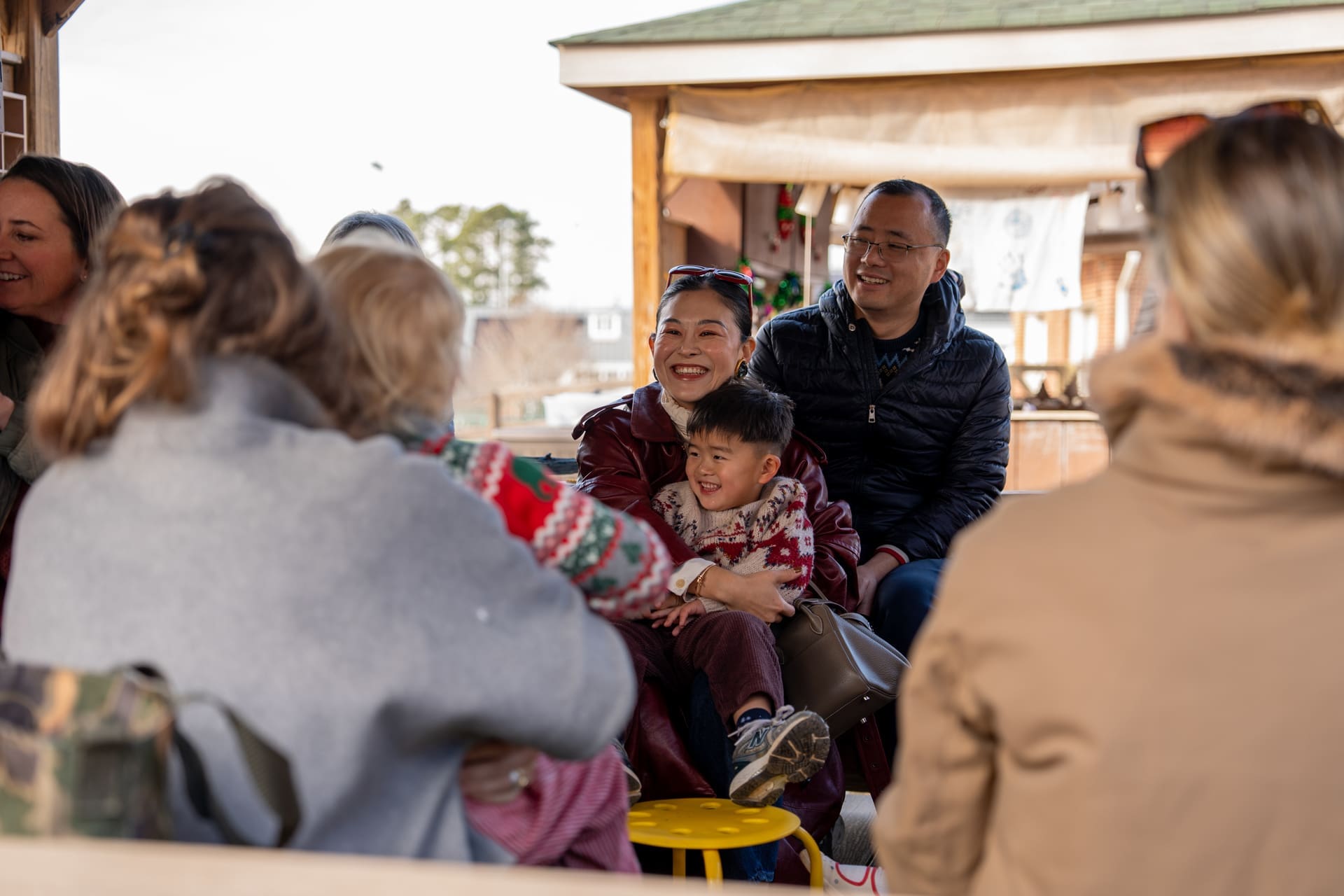 A group of adults and children sit together outdoors, smiling and talking. A woman in red holds a young boy on her lap. They appear to be enjoying a casual, friendly gathering near wooden structures.