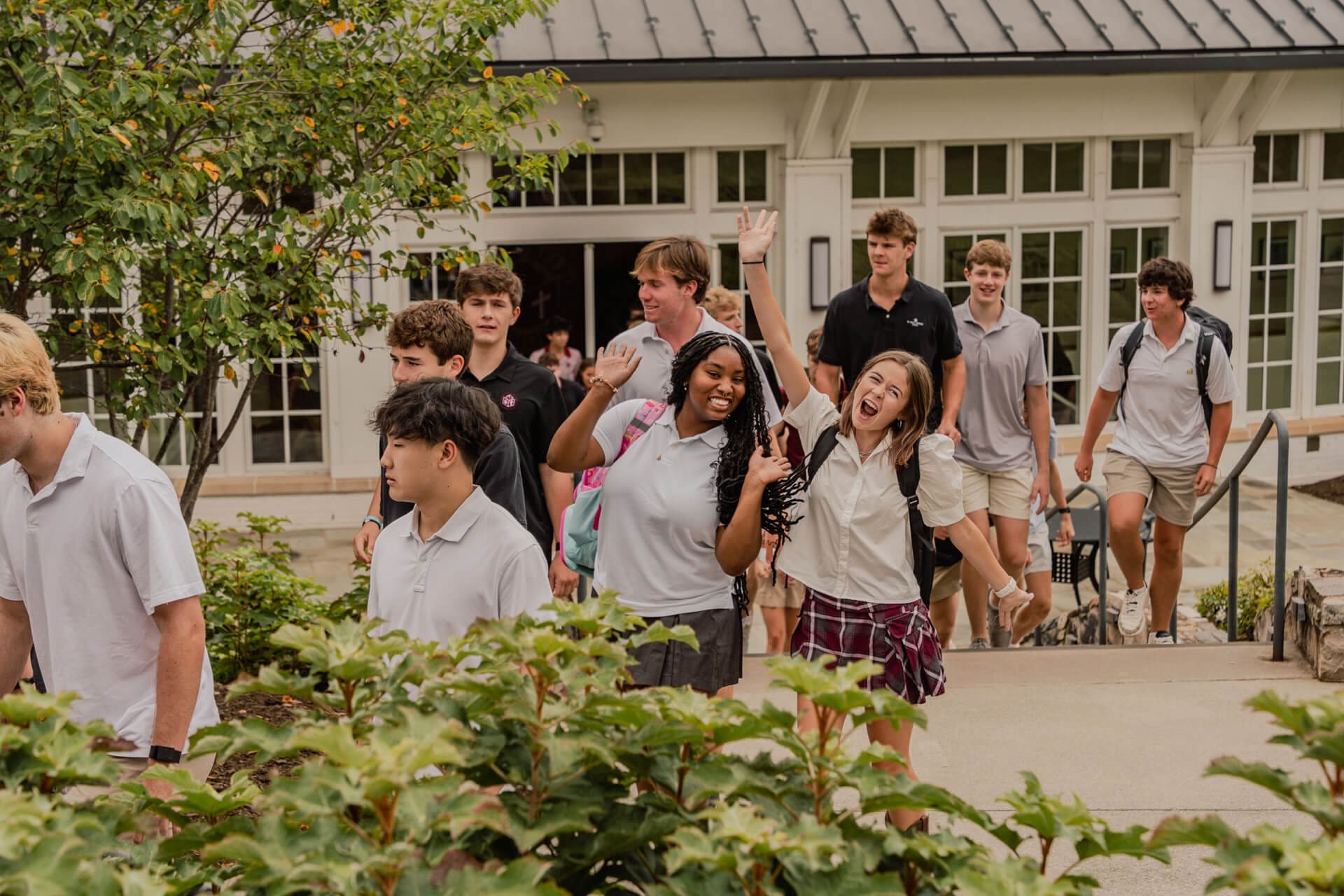 A group of high school students in uniforms walk outside a school building. Two girls in front smile and wave at the camera, while other students walk behind them. Lush green plants line the walkway.