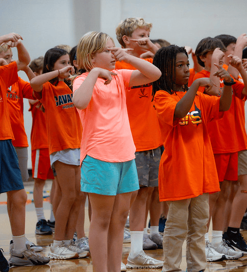 A group of children in orange shirts stand in a gymnasium, all making a hand gesture near their eyes.