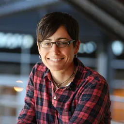 A person with short dark hair and glasses wearing a red and black plaid shirt smiles at the camera.