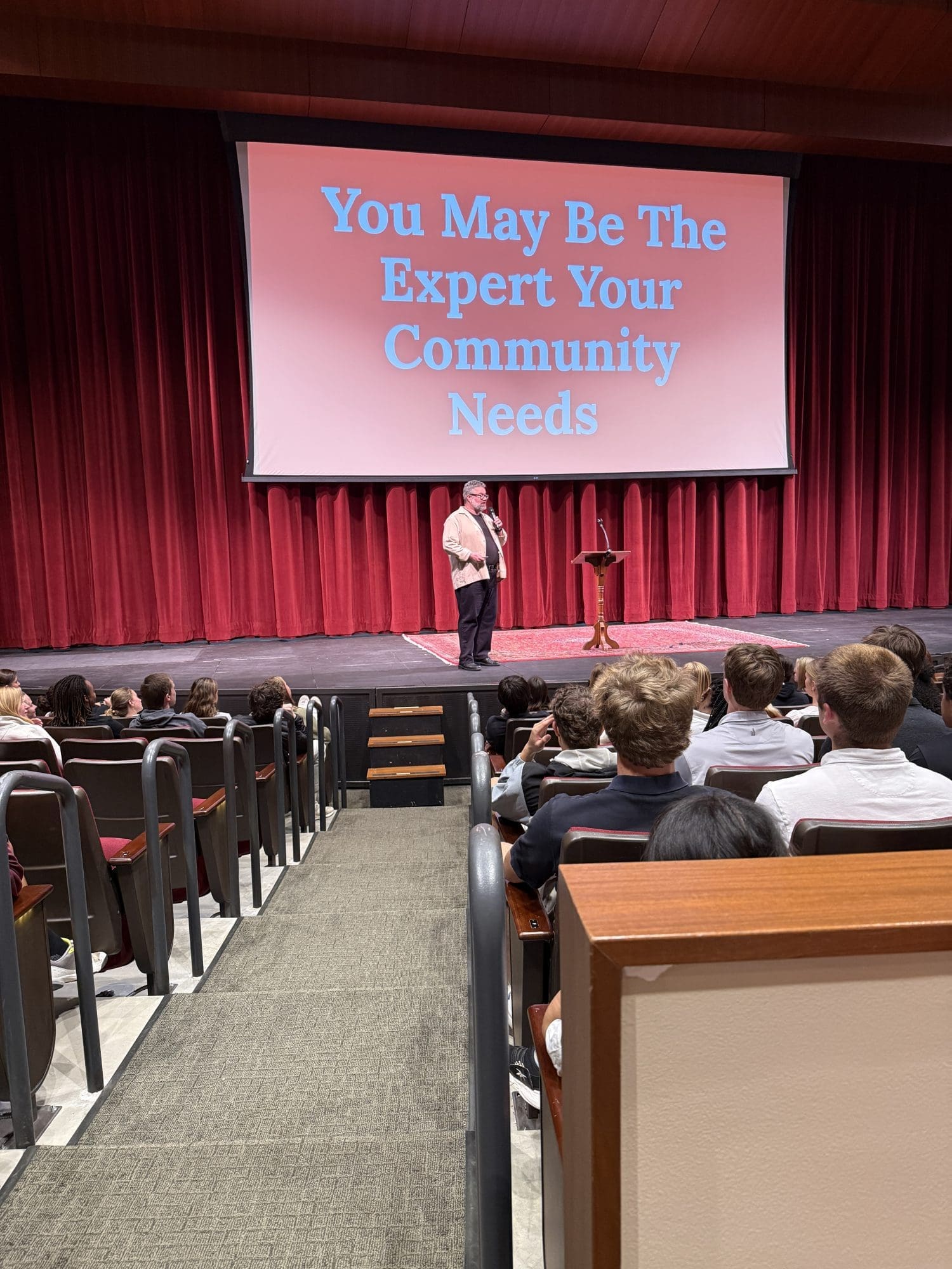 A person stands on a stage at a podium speaking to an audience, with a large screen behind them reading, You May Be The Expert Your Community Needs.
