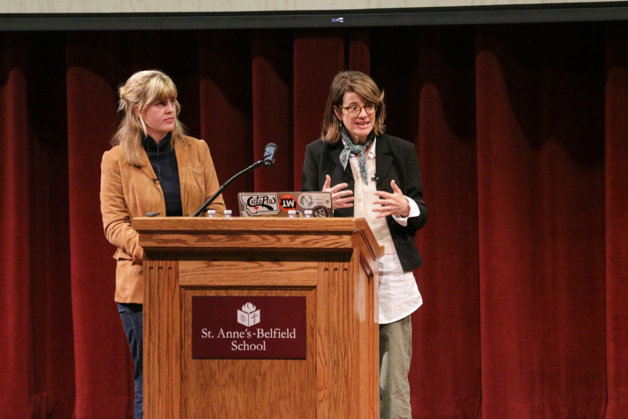 A woman stands at a wooden podium with the St. Annes-Belfield School logo, gesturing while speaking to an audience, as another woman stands beside her.
