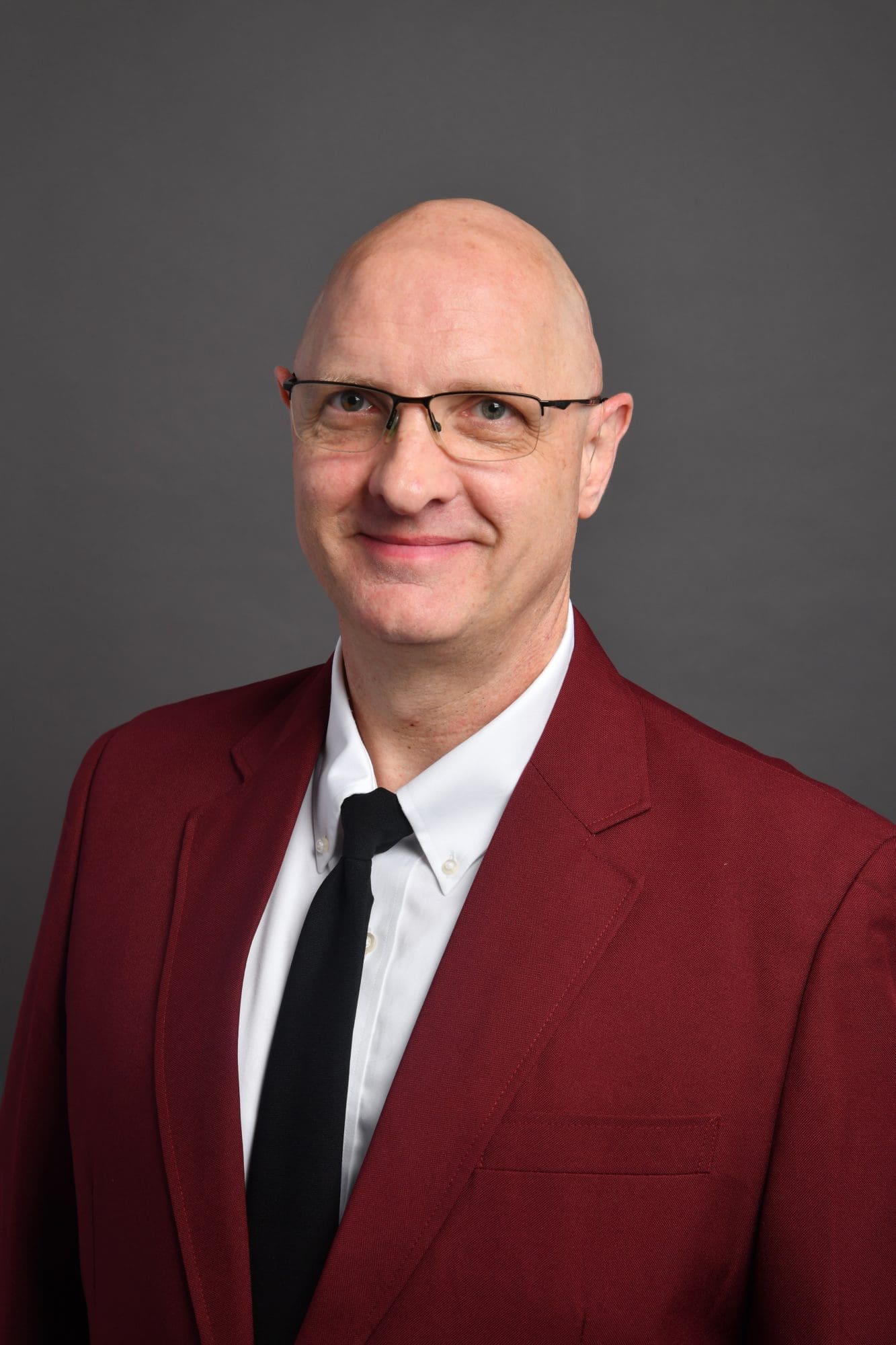 A bald man wearing glasses, a white shirt, black tie, and a burgundy suit jacket, smiling at the camera against a plain gray background.