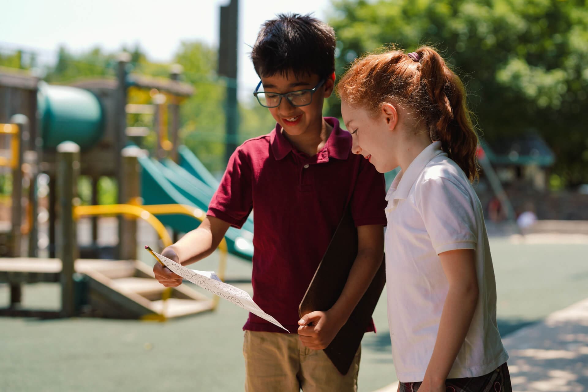 Two children stand outside near a playground, smiling and looking at a piece of paper together. One holds a pencil, and both appear engaged and happy, surrounded by greenery and playground equipment.