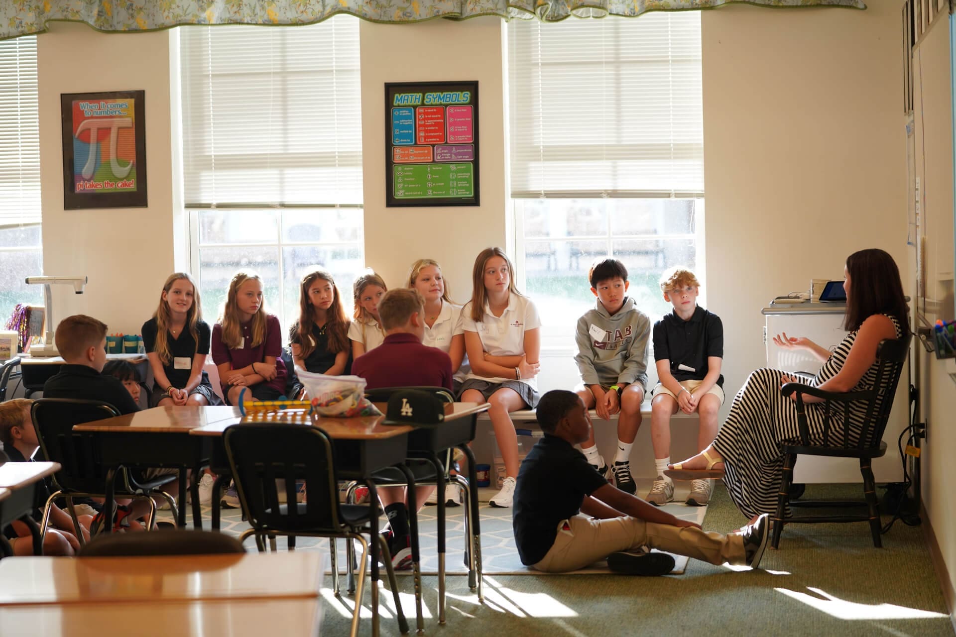 A group of students sits in a semi-circle on the floor and chairs, listening to a teacher who is seated and speaking. The classroom is well-lit with posters on the walls and large windows letting in sunlight.